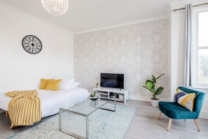 A modern living room with a bed, a TV on a white stand, a glass coffee table, a blue armchair, and a decorative clock on the wall. - ☆LUXURY APARTMENT☆BUSINESS/RELOCATIONS☆2BED (Ilford)