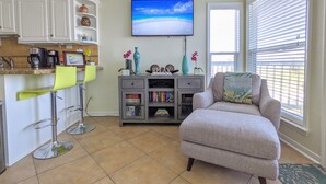 Ein Wohnzimmer mit grauem Sessel, Fernseher an der Wand und einer Küchenzeile mit Barhocker und Kaffeemaschine.