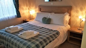 A neatly made bed with a plaid bedspread, two heart-shaped pillows, and two folded towels. There are bedside lamps on either side of the bed. - Little Fawn Felmoor Park (Morpeth)