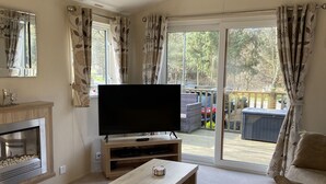 A living room with a flat-screen TV, a fireplace, a sofa, and a sliding glass door leading to an outdoor area with a deck and cars. - Little Fawn Felmoor Park (Morpeth)