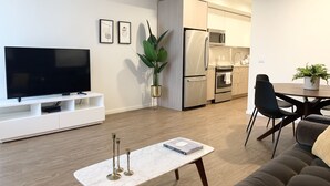 A modern living room with a flat-screen TV, a white coffee table, and a sofa. There is a dining area with a table and chairs, and a kitchen with a refrigerator and oven. - Domicile Suites at Venn on Main (Bellevue)