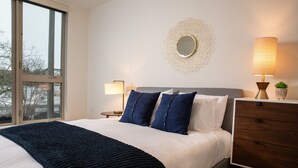 A neatly made bed with a dark blue blanket, a bedside table with a lamp, and a decorative mirror on the wall. - Domicile Suites at Venn on Main (Bellevue)