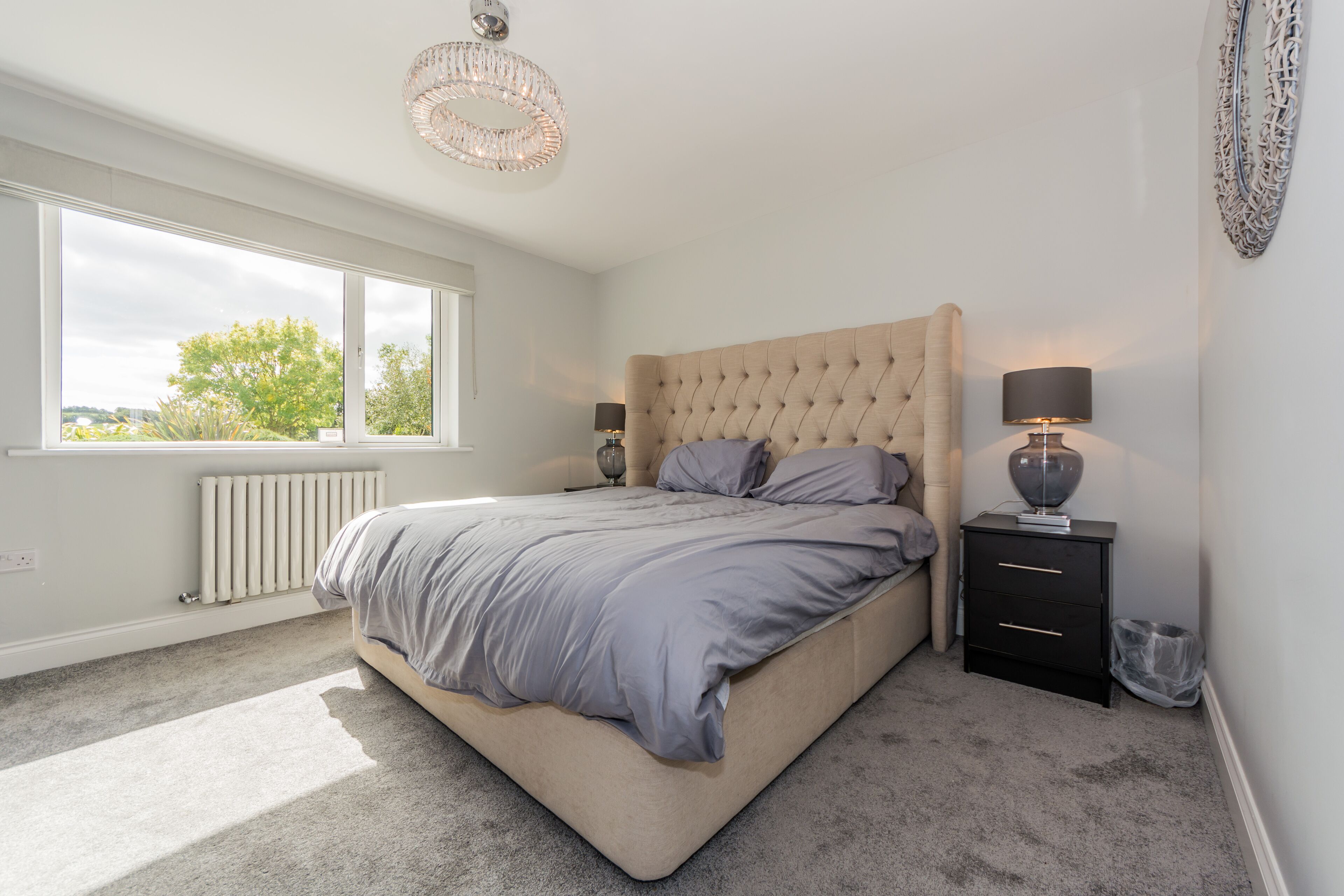 A bedroom with a large bed, two bedside tables, a radiator, and a window with a view of trees.