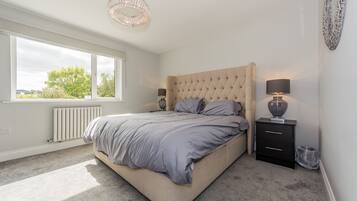 A bedroom with a large bed, two bedside tables, a radiator, and a window with a view of trees.