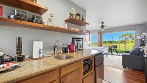 Cocina moderna con encimeras de granito, armarios de madera y horno empotrado. Cuenta con un comedor con sofá gris y vistas al exterior.