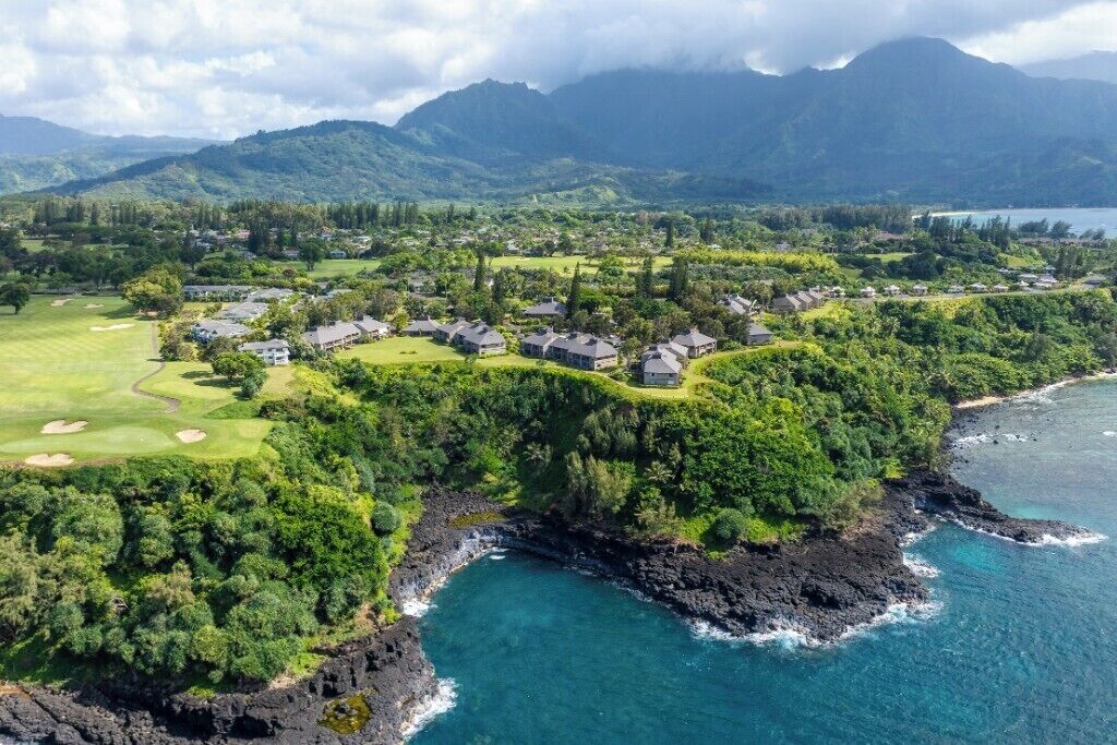 An aerial view of a coastal resort with houses arranged in a crescent shape along the shoreline, a golf course, and lush greenery.