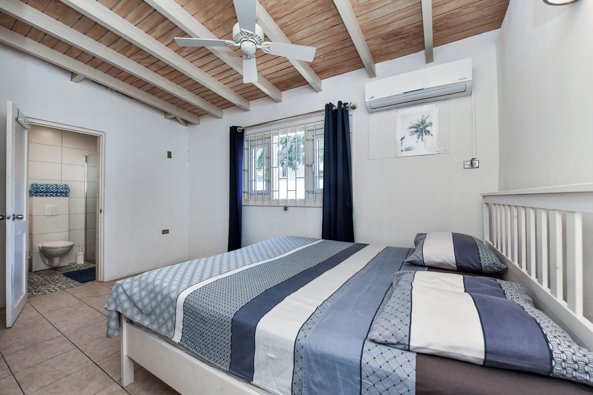 Une chambre à coucher comprenant un lit, un ventilateur de plafond, une fenêtre avec des rideaux et un appareil de climatisation.