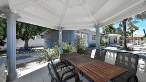 Eine Terrasse im Freien mit einem Holztisch und -stühlen, umgeben von Palmen und einer weißen Laube.