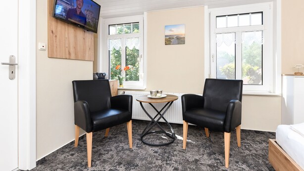 Une pièce comprenant deux fauteuils en cuir noir, une petite table ronde, un téléviseur fixé au mur et un lit avec une tête de lit en bois.