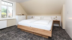 Une chambre avec un plafond en pente, une grande fenêtre, un lit avec une literie blanche, une table de chevet avec une lampe et une petite armoire.