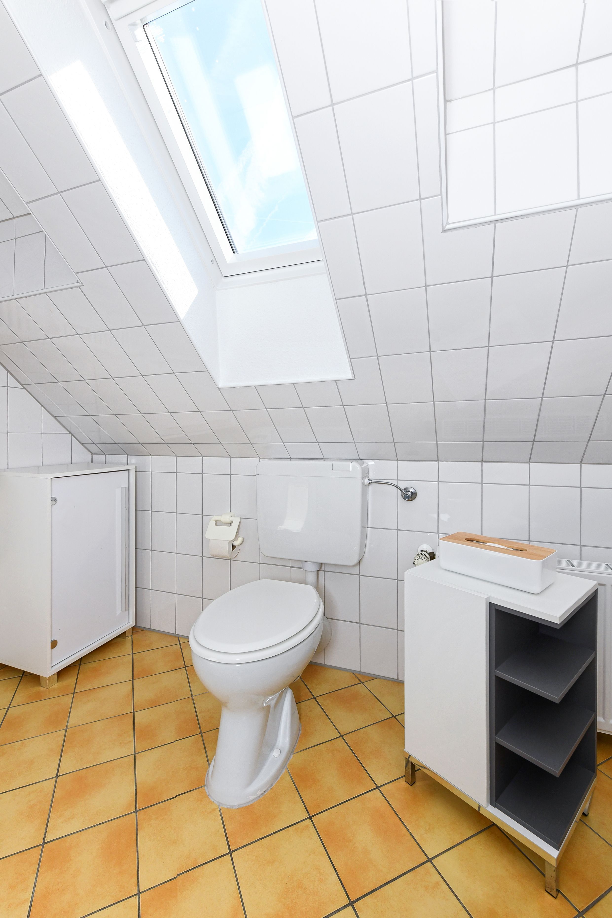 Uma casa de banho moderna com um claraboia, azulejos brancos, uma sanita e um armário com prateleiras.