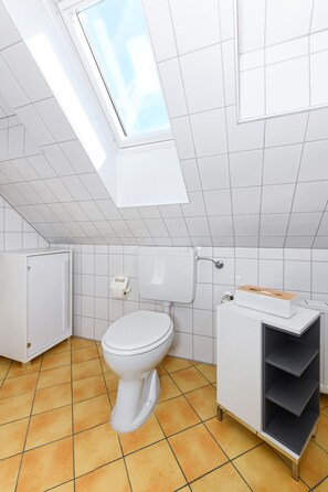 Et moderne badeværelse med ovenlys, hvide fliser, toilet og et skab med hylder.