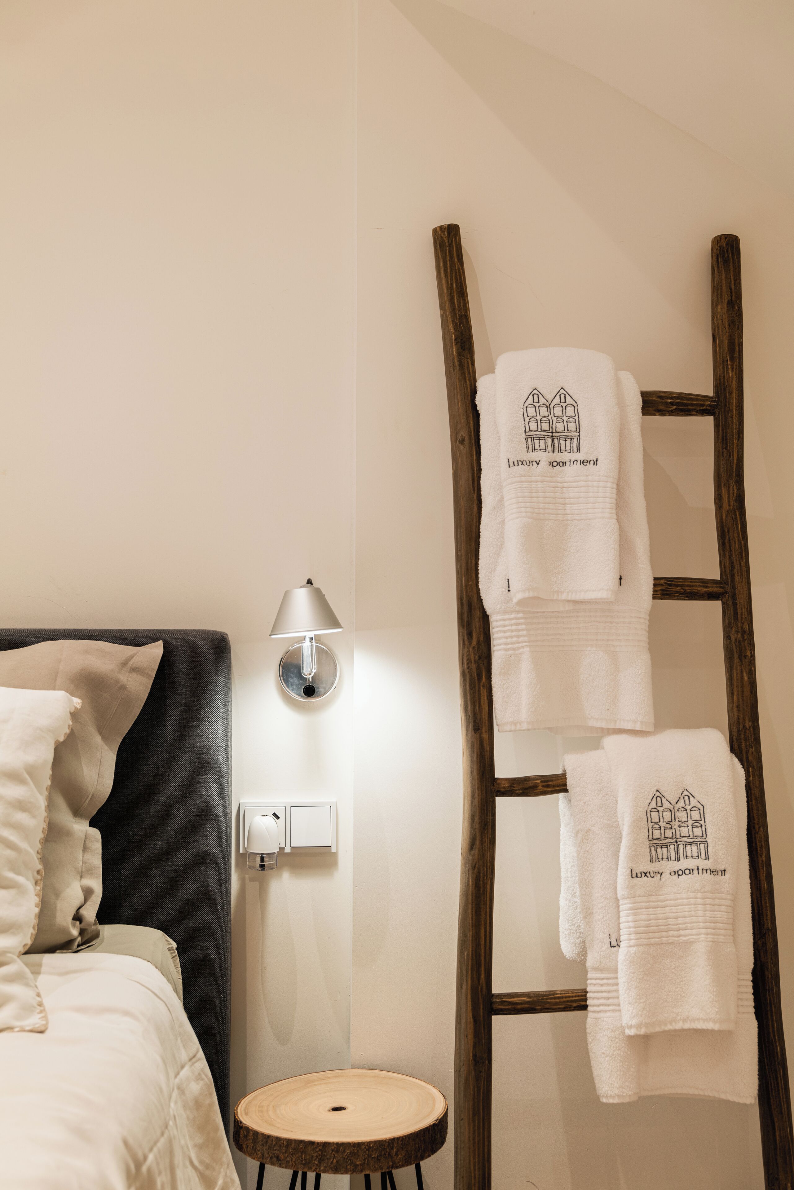 Una cama bien ordenada con cabecera, una lámpara de noche y un pequeño taburete de madera.