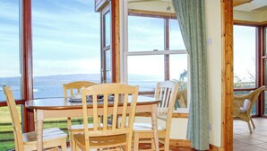Un espace repas comprenant une table et des chaises, de grandes fenêtres donnant sur l’extérieur et un sol carrelé.