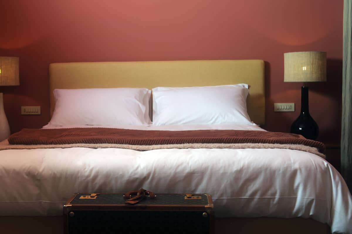 Een netjes opgemaakt bed met witte lakens, een bruine plaid en een kleine koffer op het voeteneinde.
