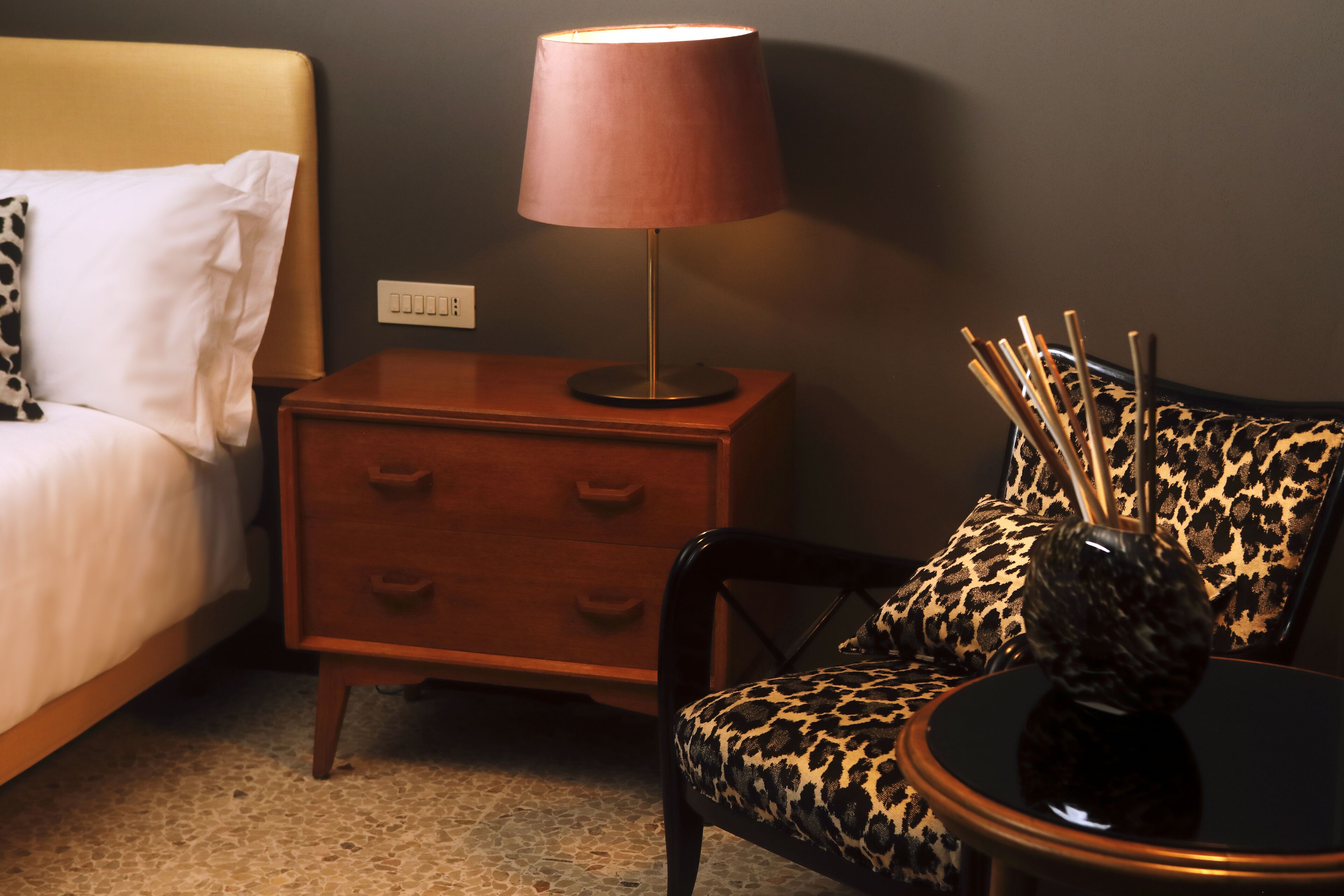 A hotel room with a bed, a nightstand with a lamp, a chair with a leopard print cushion, and a small table with a vase.