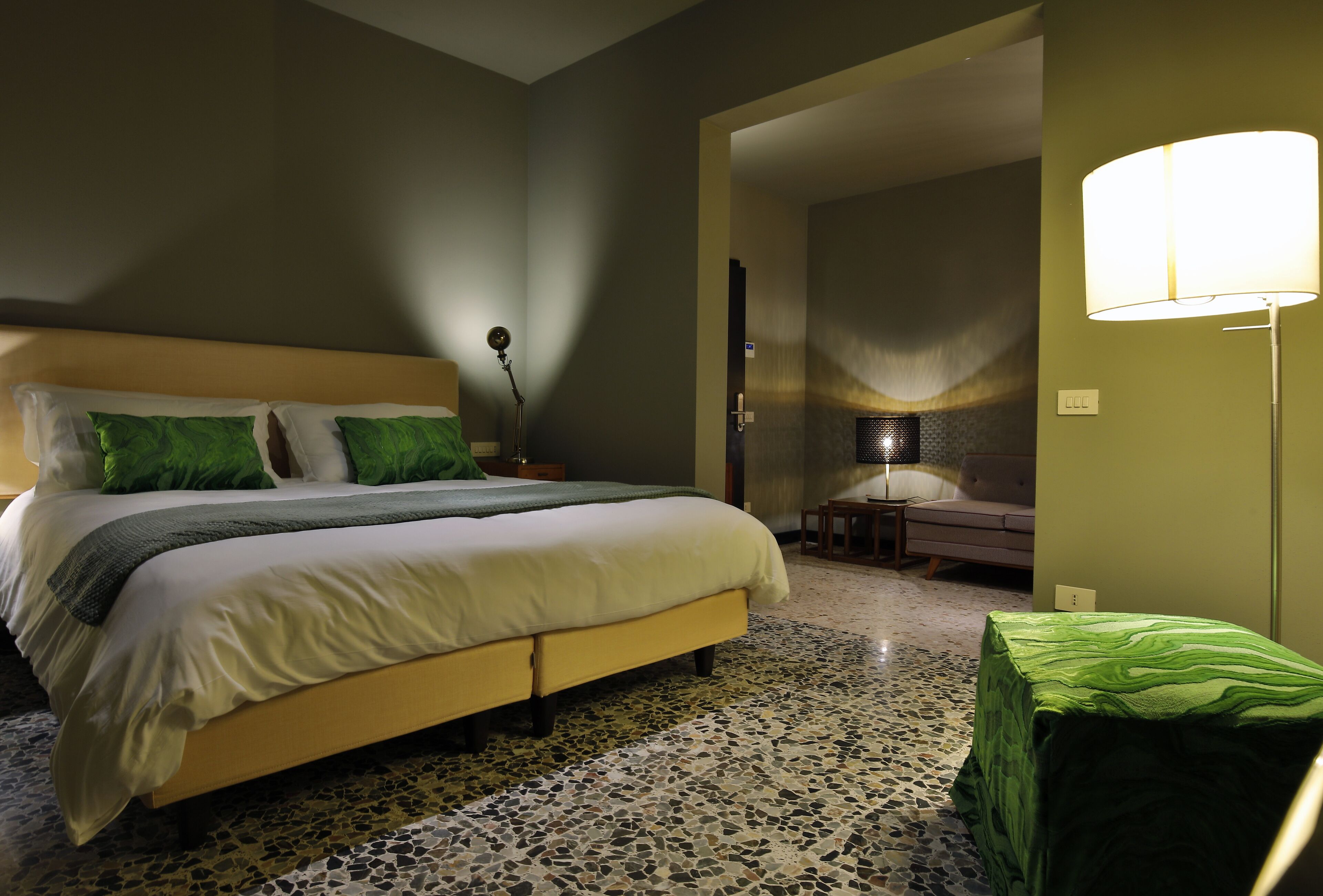 Ein Schlafzimmer mit einem Bett, grünen Kissen, einem Nachttisch, einer Lampe und einem mit Grün bezogenen Hocker.