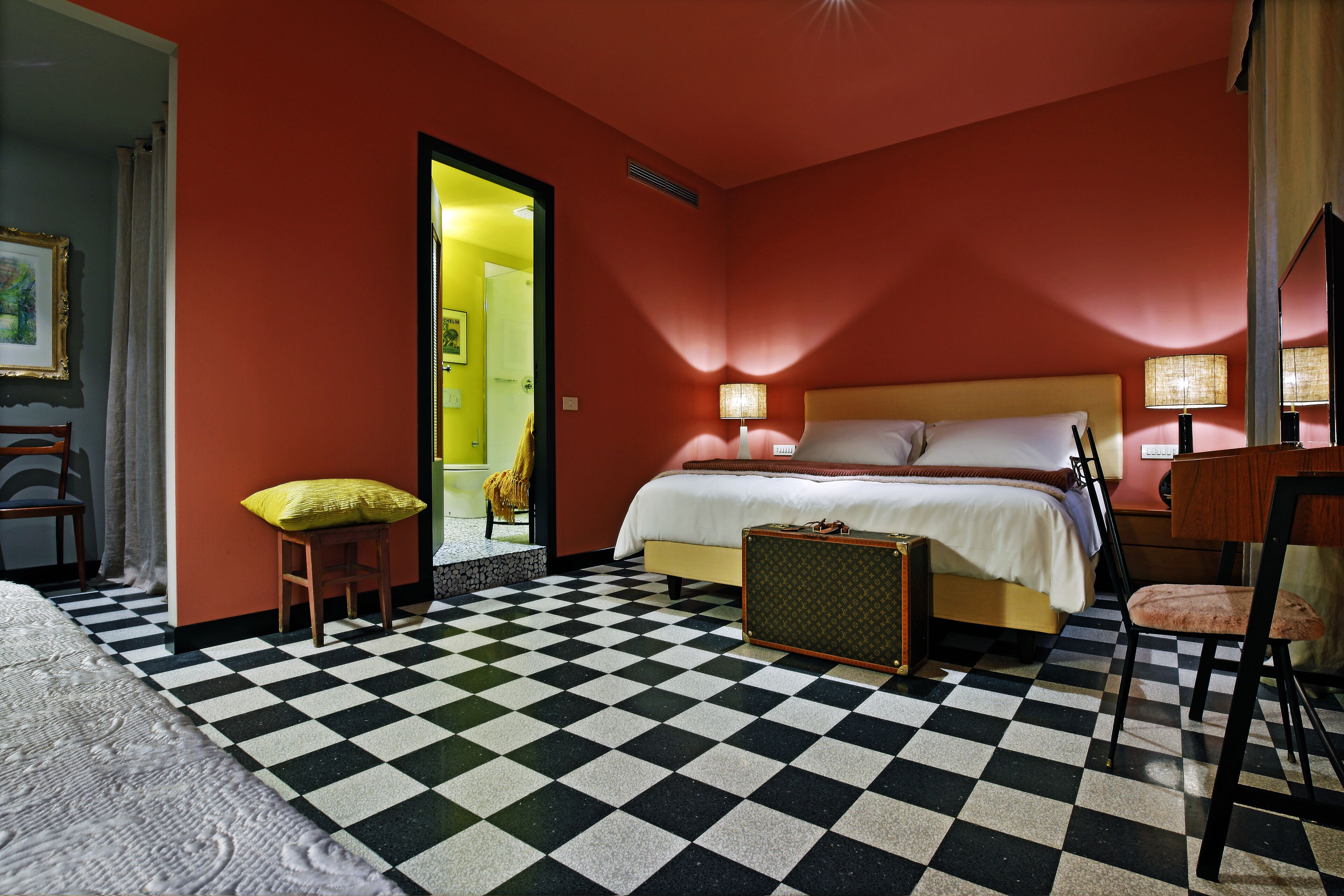 A hotel room with a checkered floor, a bed with white linens, a wooden nightstand, and a suitcase.
