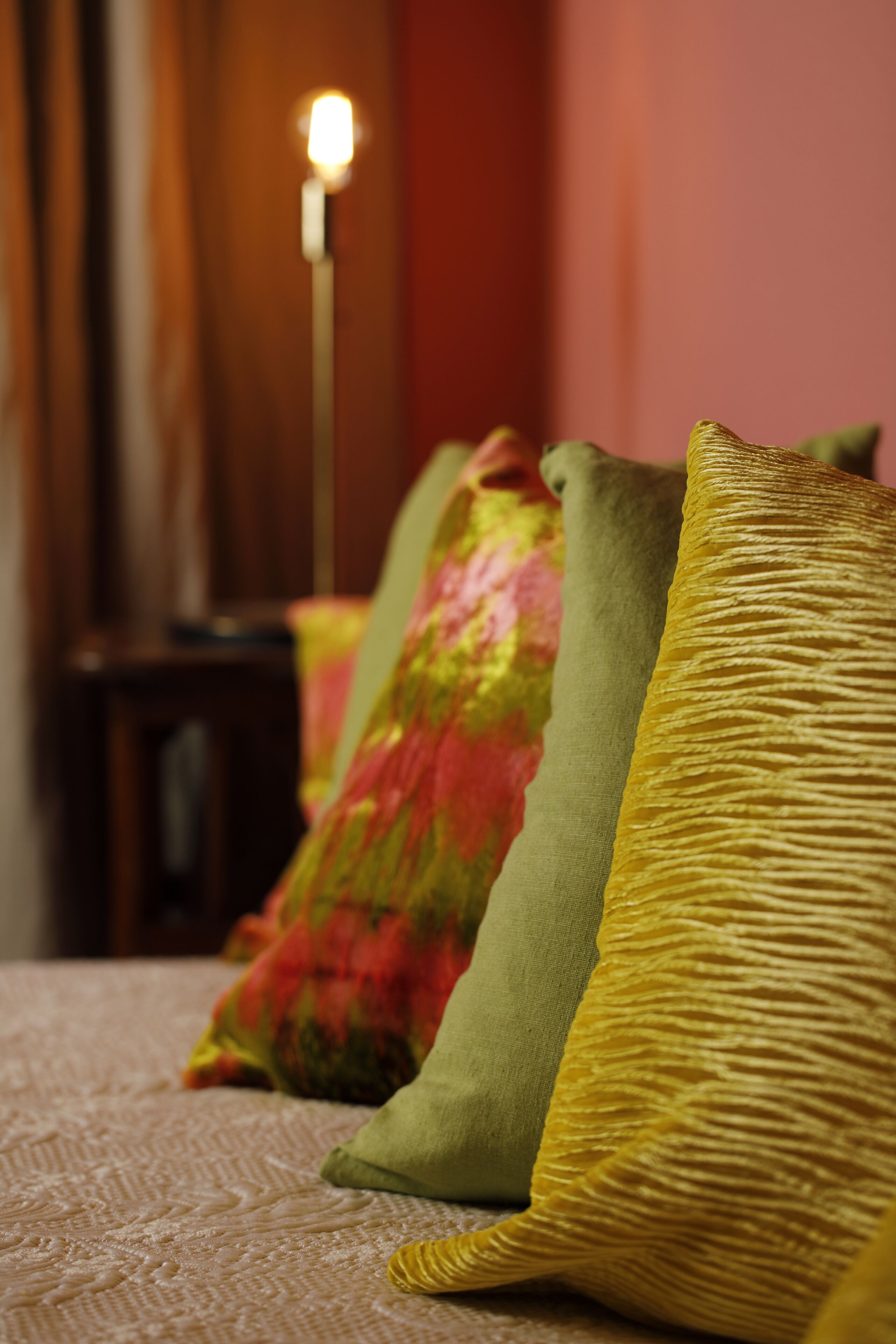 Ein Bett mit Kissen in verschiedenen Farben und Texturen, ein Nachttisch und eine Lampe im Hintergrund.