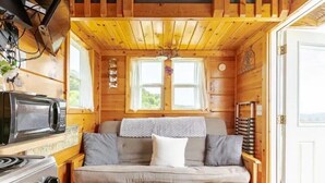 A cozy cabin interior with a wooden ceiling, a sofa with cushions, a microwave, a stove, and a window with curtains.