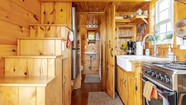 A wooden cabin interior with a kitchen area, wooden staircase, and a visible bathroom.