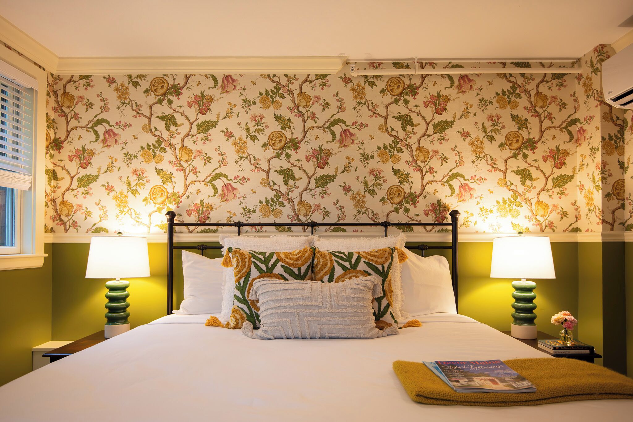 Una cama con almohadas de estampado floral, dos lámparas de noche y una revista sobre la cama.