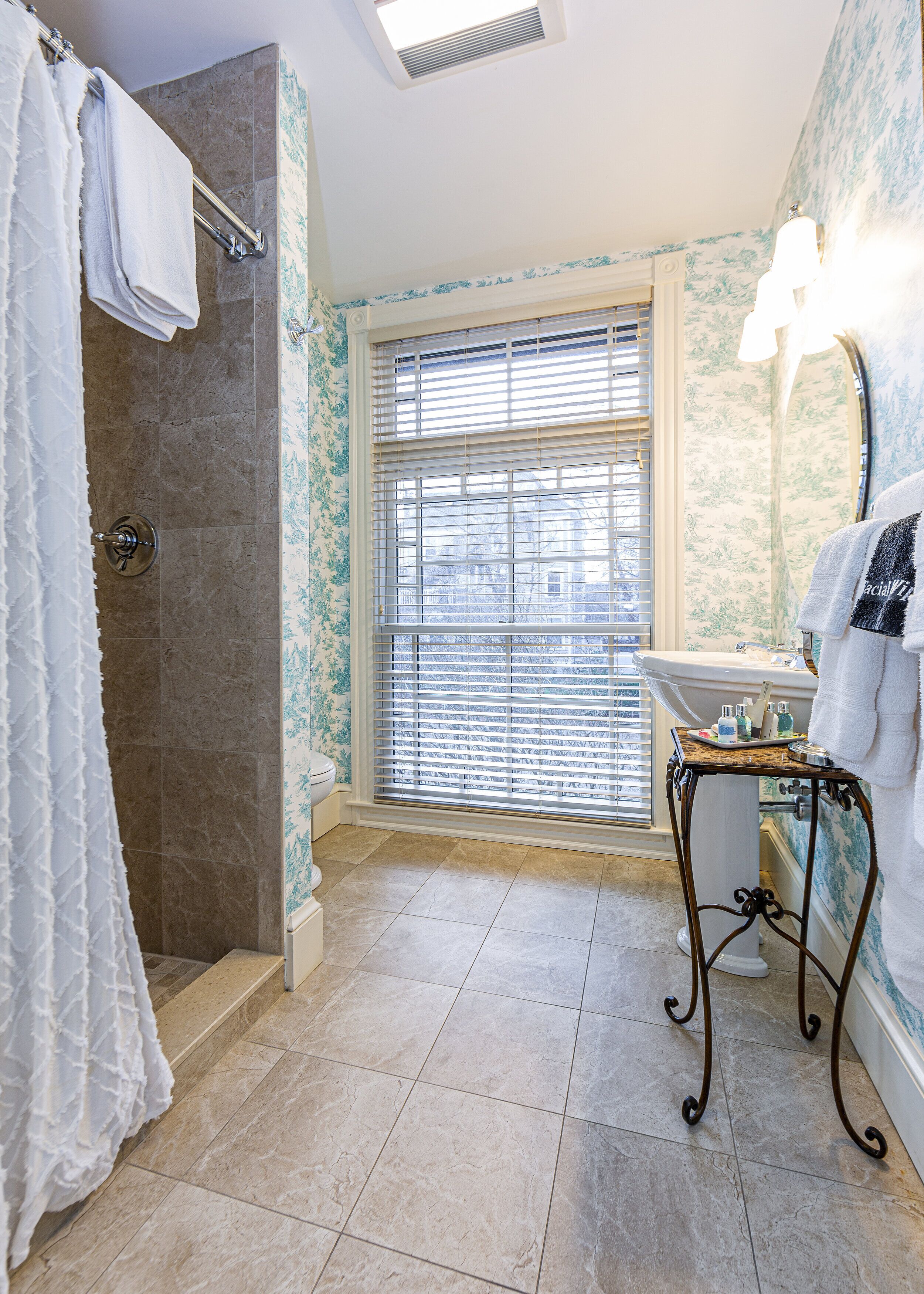A bathroom with a shower, a vanity with a mirror, and a window with blinds.