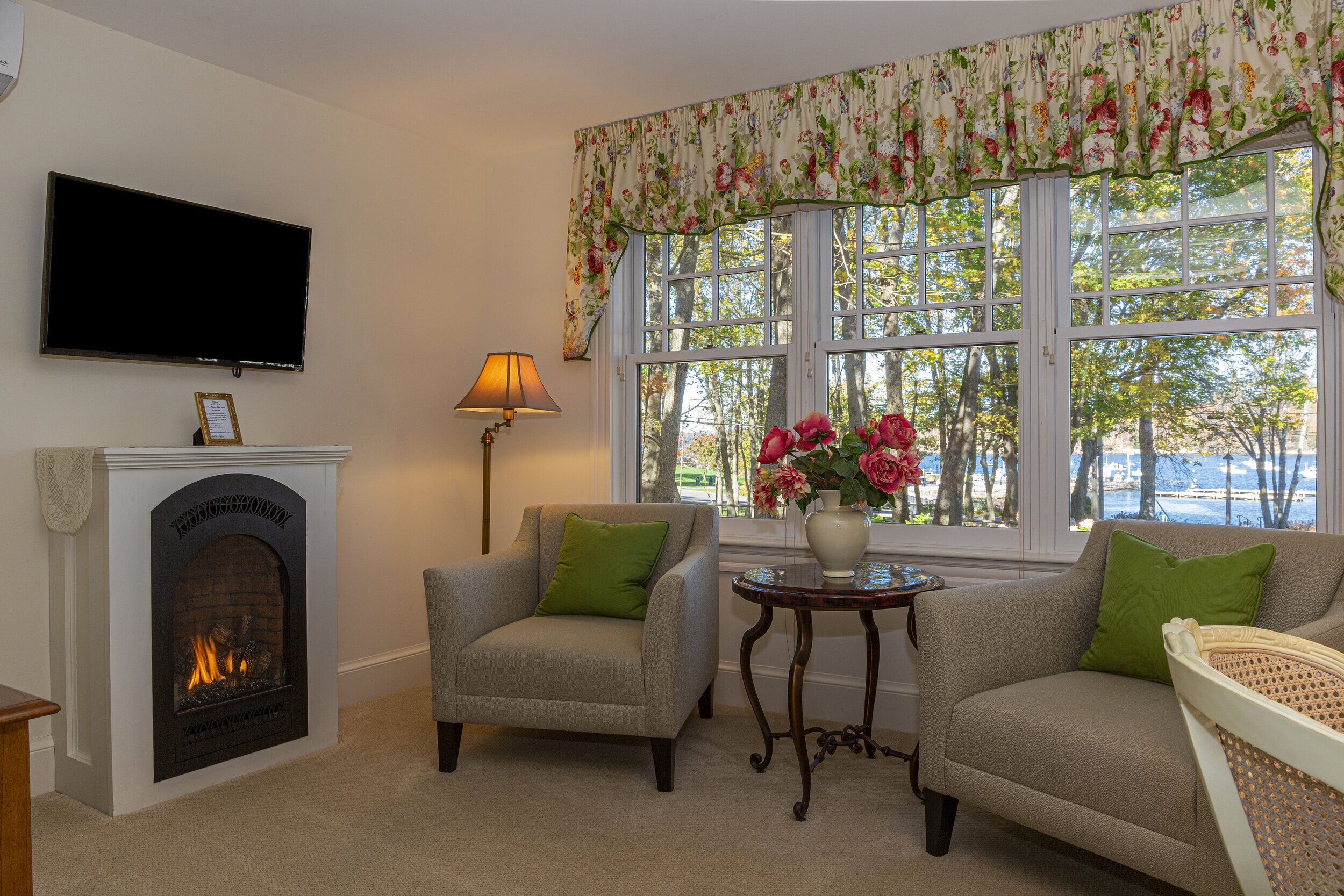 Ein Wohnzimmer mit Kamin, einem an der Wand befestigten Fernseher, einer Couch, einem kleinen Tisch mit Blumenvase und einem Fenster mit blumigem Vorhang.