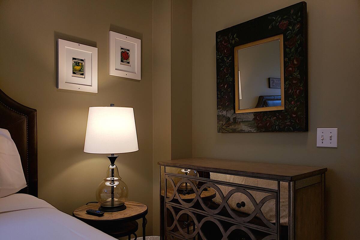 A hotel room with a bed, a nightstand with a lamp, a dresser, and two framed pictures on the wall.