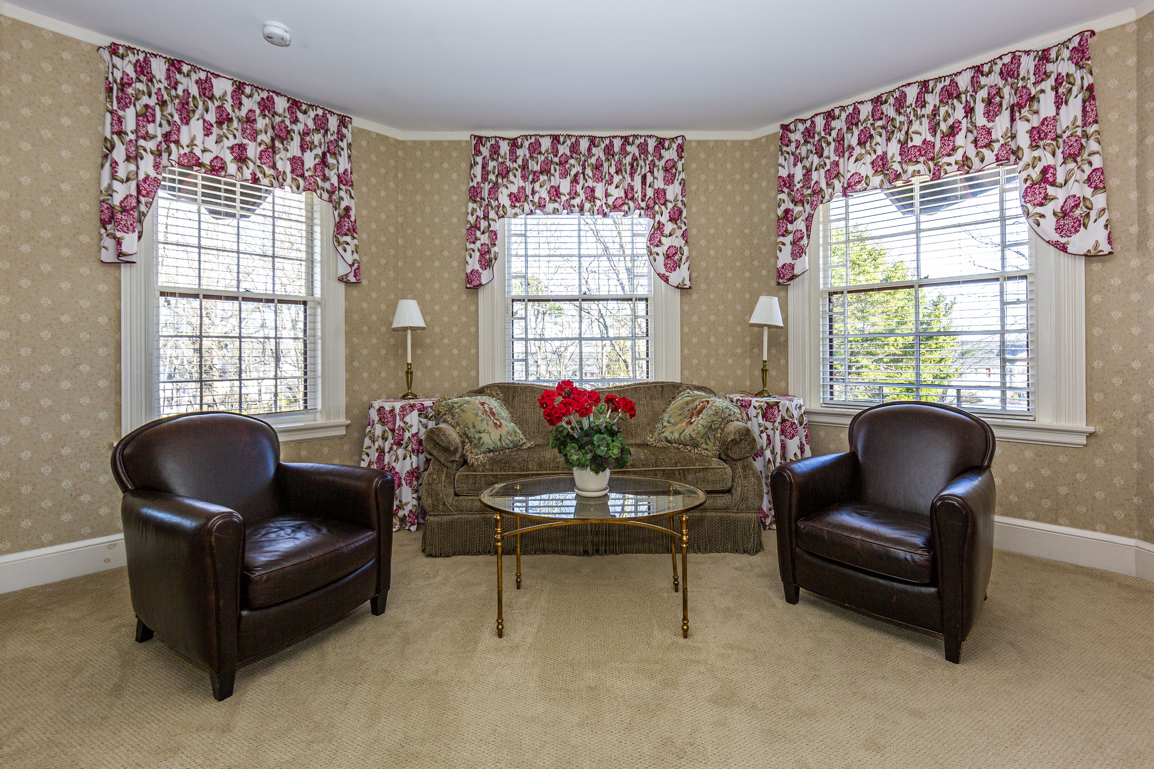 A room with a brown leather sofa, two leather armchairs, a glass coffee table with a floral vase, and floral curtains on the windows.