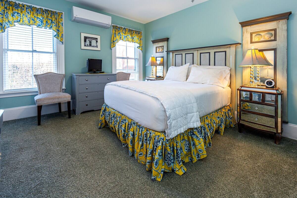 A bedroom with a large bed, a dresser, a chair, a nightstand with a lamp, and a window with curtains.