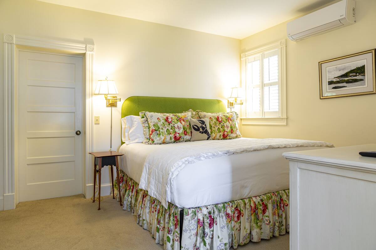 A bedroom with a green upholstered headboard, floral bedding, a bedside table with a lamp, and a framed picture on the wall.