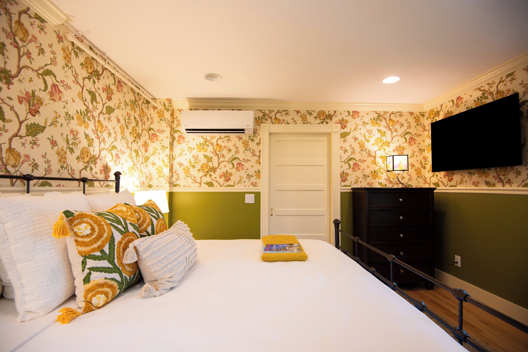 A bedroom with floral wallpaper, a bed with pillows, a bedside table, a dresser, an air conditioning unit, a television, and a door.