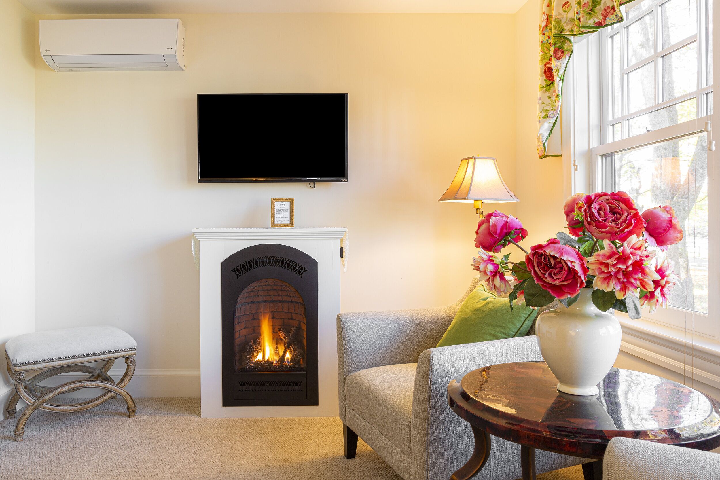 A living room with a fireplace, a television mounted on the wall, a comfortable sofa, a small table with a vase of flowers, and a window with curtains.