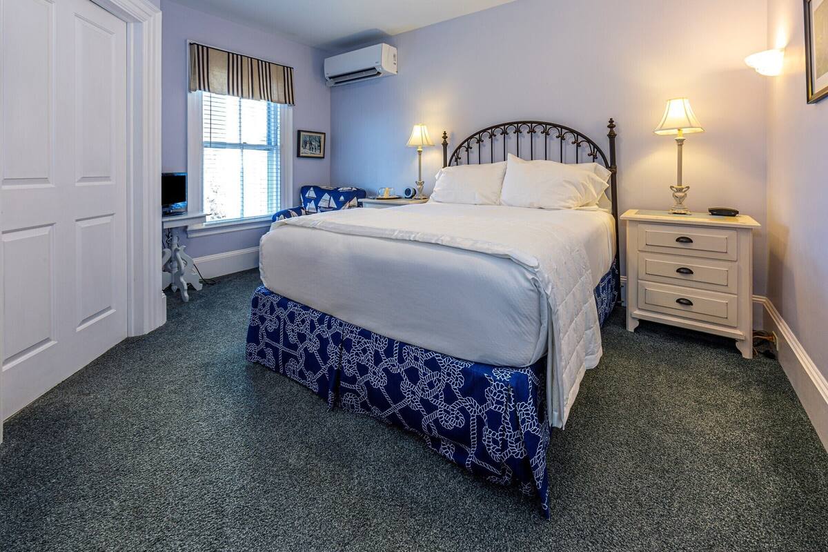 A bedroom with a bed, bedside tables, a lamp, a window with curtains, and an air conditioning unit.