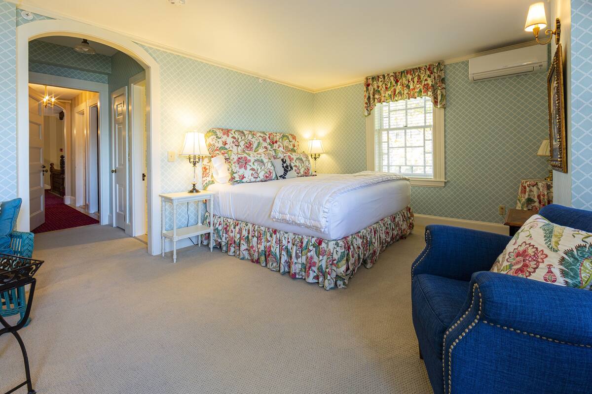 A bedroom with a floral bedspread, a white headboard, and a window with curtains. There is a chair with a floral cushion and a small table with a lamp.