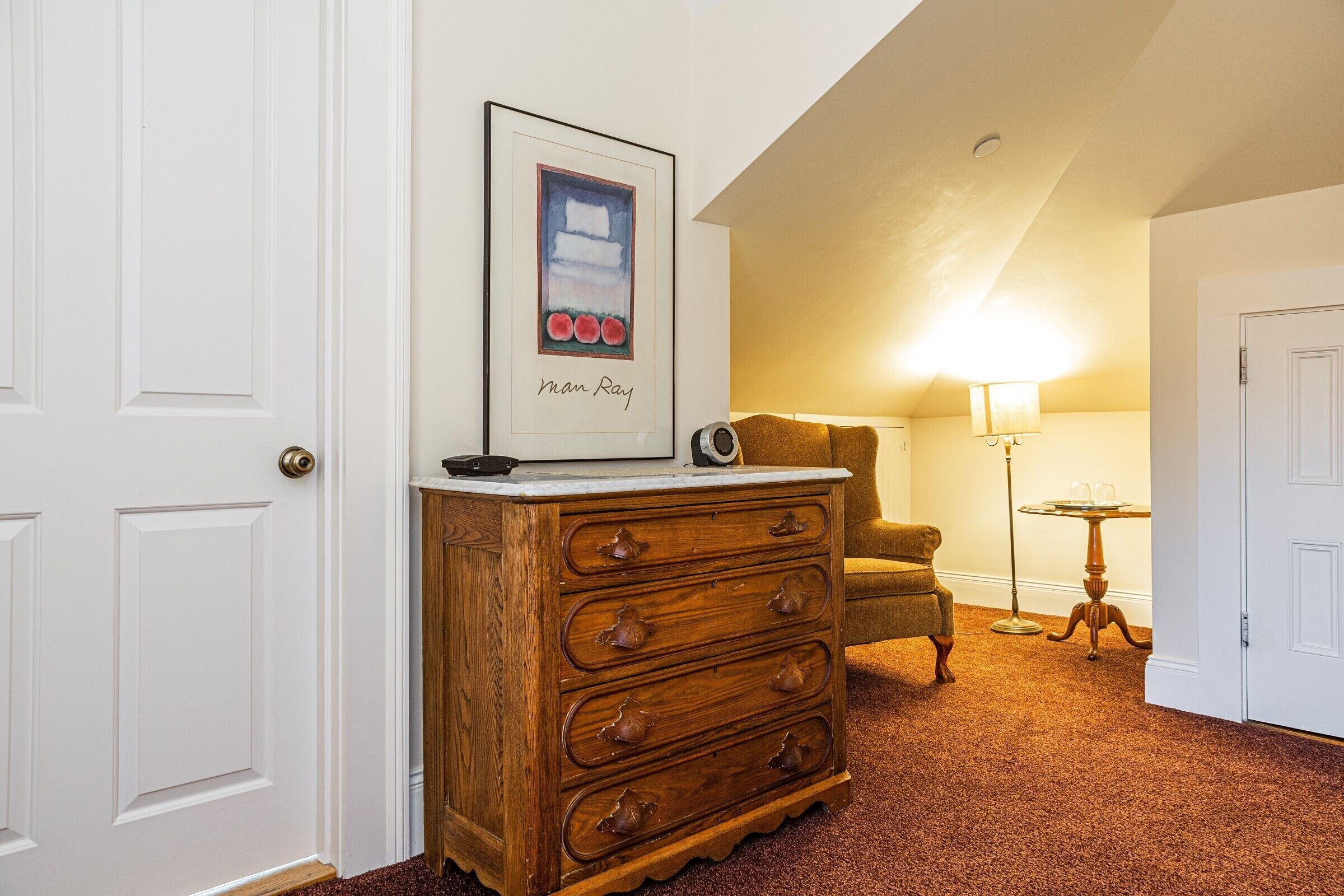 A room with a wooden dresser, a framed artwork, a lamp, a chair, and a door.