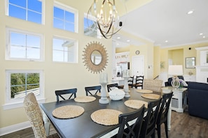 Uma sala de jantar com uma mesa grande, cadeiras e um candelabro. Há janelas com vista para áreas verdes lá fora.