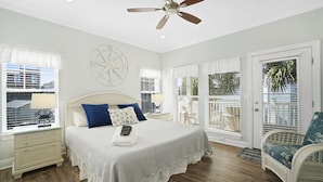Une chambre à coucher comprenant un lit, des tables de chevet, un ventilateur de plafond, une fenêtre avec des stores, une porte et un fauteuil en osier.