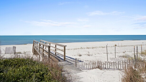 Une promenade en bois mène à une plage de sable fin, sous un ciel bleu et dégagé.