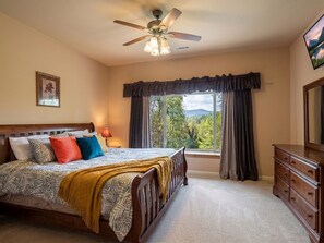A bedroom with a bed, a dresser, a ceiling fan, and a window with a view of trees and mountains.