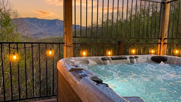 Un jacuzzi con cubierta de madera y baranda de metal, rodeado por un paisaje natural con montañas al fondo.