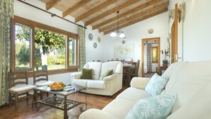 A living room with a white sofa, a coffee table with oranges, and a dining area with wooden furniture. - VILLA FINQUITA - RUSTIC STAY (Puerto Pollensa)