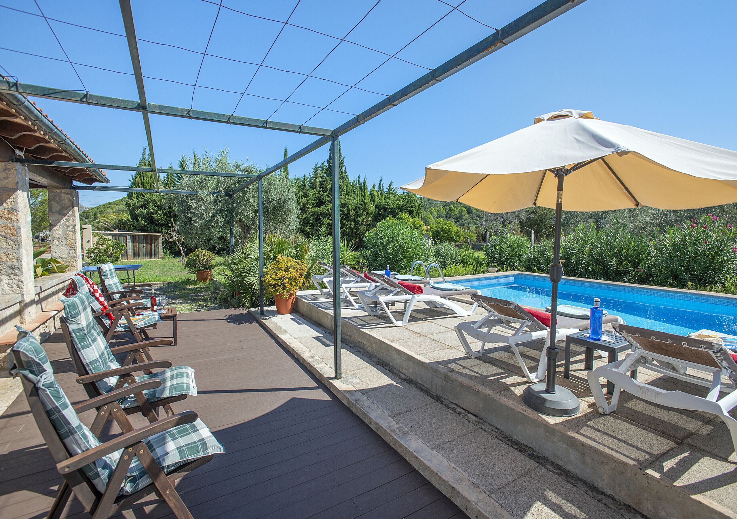 A poolside area with lounge chairs and a large umbrella, surrounded by greenery and a clear blue sky.