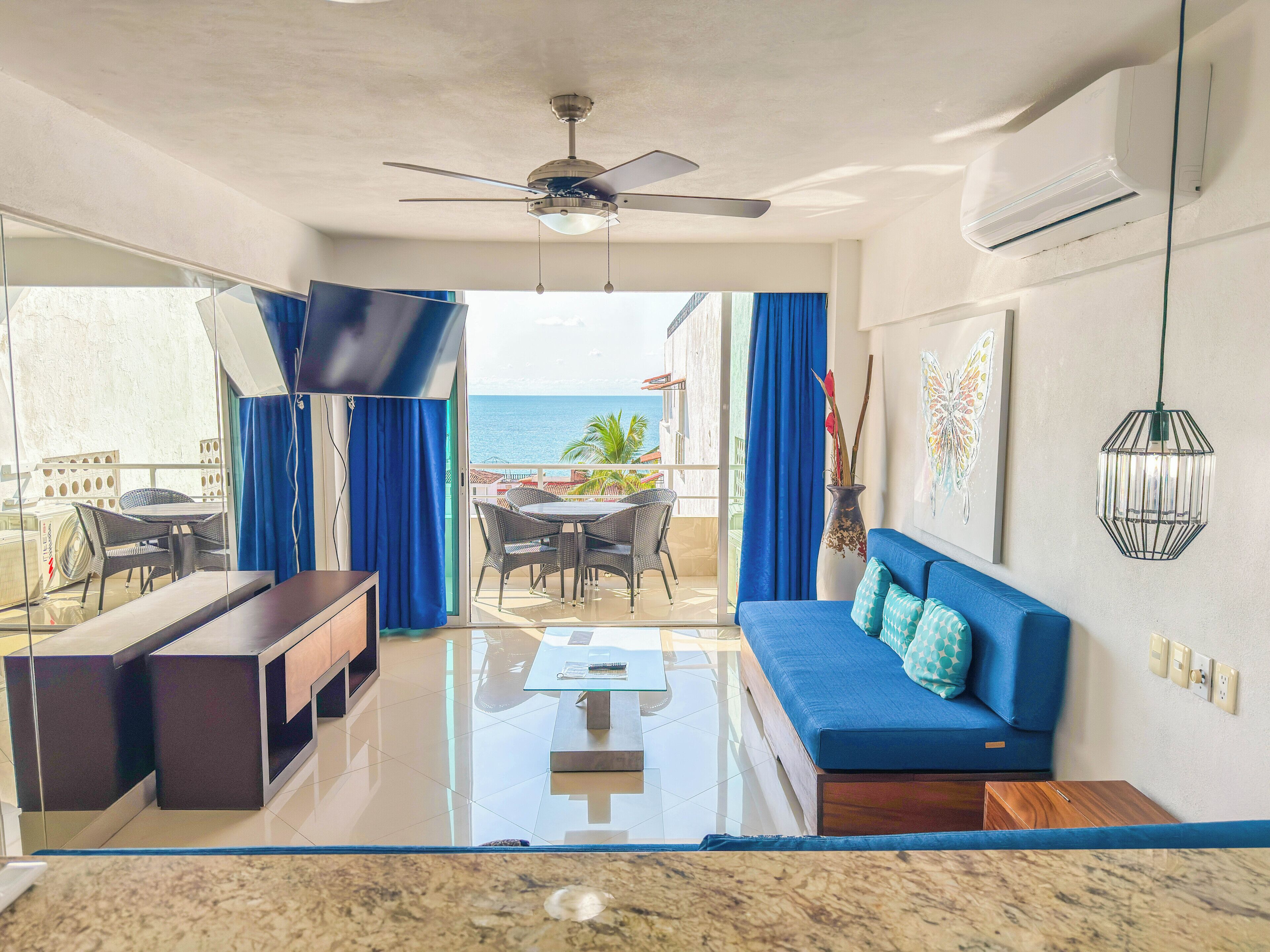 A modern living room with a sofa, coffee table, and chairs, a ceiling fan, and a view of the ocean.