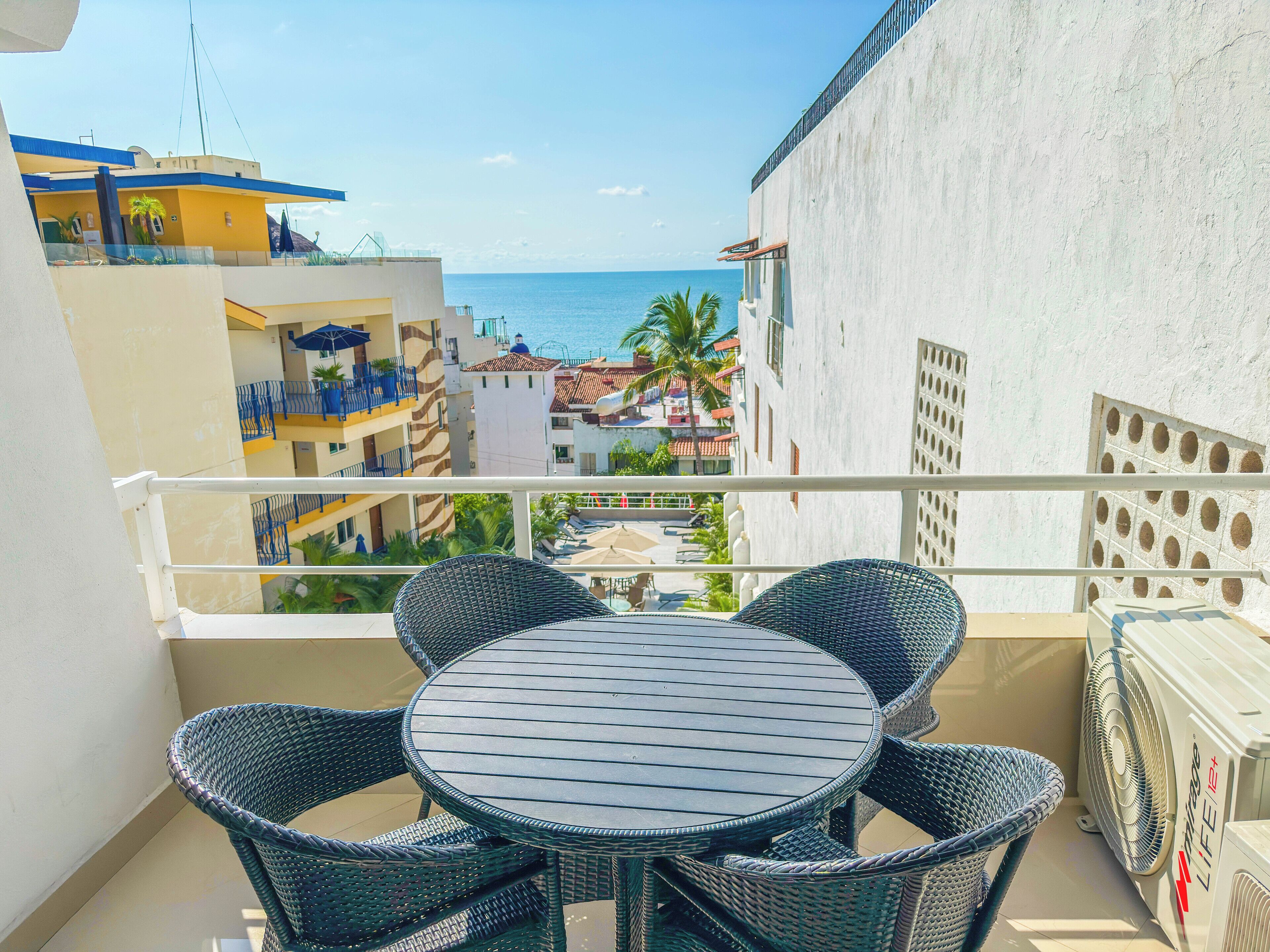 A balcony with wicker chairs and a table, overlooking a coastal area with buildings and a view of the sea.