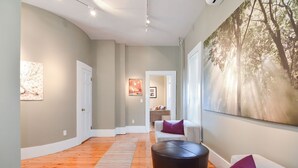 A modern living room with a wooden floor, a large painting of trees, and a sofa with purple cushions.