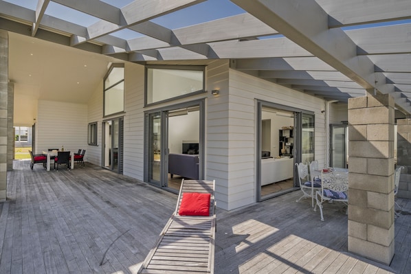 A modern outdoor patio with a wooden deck, a lounge chair with a red cushion, and a dining area with a table and chairs.