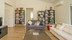 Un salon spacieux avec un canapé gris, un canapé blanc et une table basse. On y trouve deux bibliothèques remplies de livres et d’objets décoratifs. Un téléviseur est posé sur un meuble à gauche.