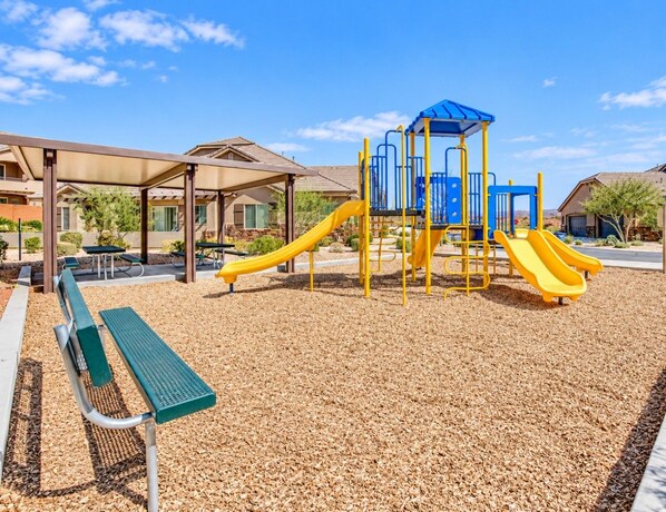 A playground with a slide and climbing structures, surrounded by a covered seating area and a building in the background.